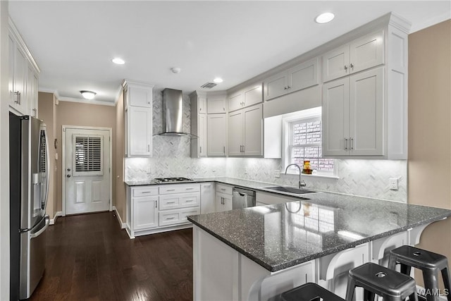 kitchen with appliances with stainless steel finishes, white cabinetry, a sink, a peninsula, and wall chimney exhaust hood