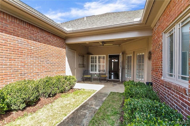 entrance to property featuring a ceiling fan, brick siding, a patio, and roof with shingles