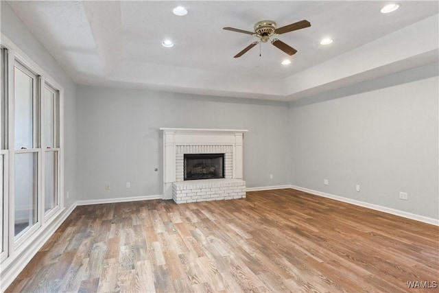 unfurnished living room featuring wood finished floors, baseboards, a tray ceiling, ceiling fan, and a brick fireplace