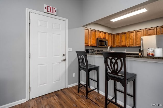 kitchen with dark countertops, dark wood-type flooring, a kitchen bar, brown cabinetry, and stainless steel appliances