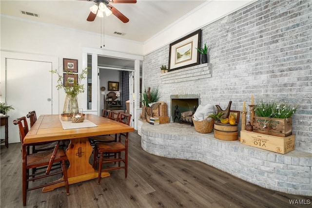 dining room featuring crown molding, ceiling fan, wood-type flooring, brick wall, and a brick fireplace