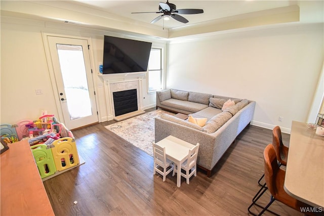 living room with dark wood-style flooring, a fireplace, a raised ceiling, ceiling fan, and baseboards