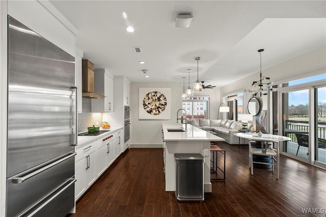 kitchen with wall chimney range hood, sink, appliances with stainless steel finishes, white cabinets, and a center island with sink