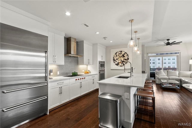 kitchen featuring an island with sink, sink, white cabinets, stainless steel appliances, and wall chimney exhaust hood