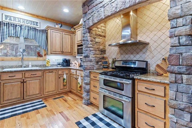 kitchen with light stone counters, stainless steel appliances, sink, wall chimney range hood, and light hardwood / wood-style flooring