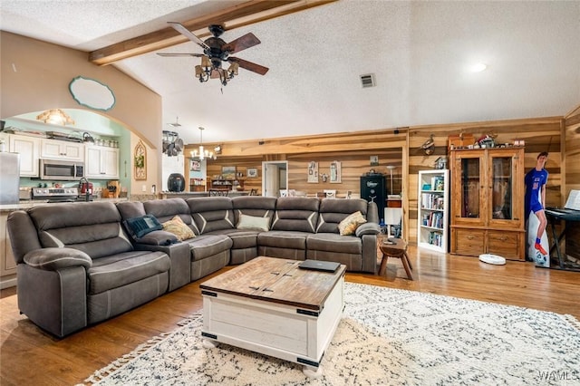 living room featuring ceiling fan with notable chandelier, wooden walls, vaulted ceiling with beams, light wood-type flooring, and a textured ceiling