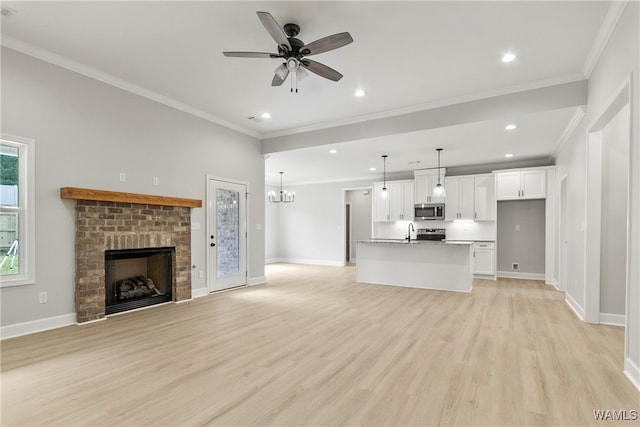 unfurnished living room featuring ceiling fan with notable chandelier, sink, crown molding, a brick fireplace, and light hardwood / wood-style flooring