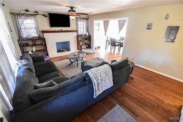 living room with a brick fireplace, ceiling fan, baseboards, and wood finished floors