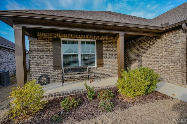 entrance to property featuring a porch, brick siding, roof with shingles, and central air condition unit