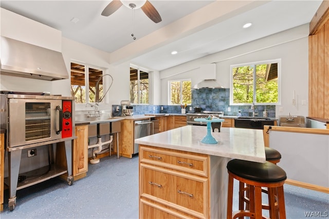 kitchen featuring appliances with stainless steel finishes, tasteful backsplash, wall chimney exhaust hood, ceiling fan, and a kitchen island