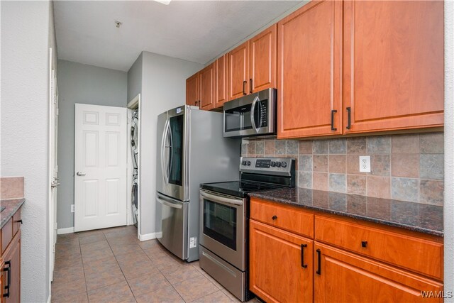 kitchen featuring backsplash, dark stone counters, stacked washer and clothes dryer, light tile patterned floors, and appliances with stainless steel finishes