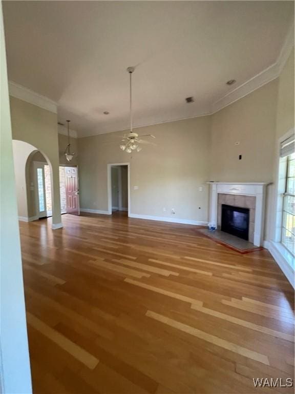 unfurnished living room featuring ceiling fan, a tiled fireplace, baseboards, wood finished floors, and ornamental molding