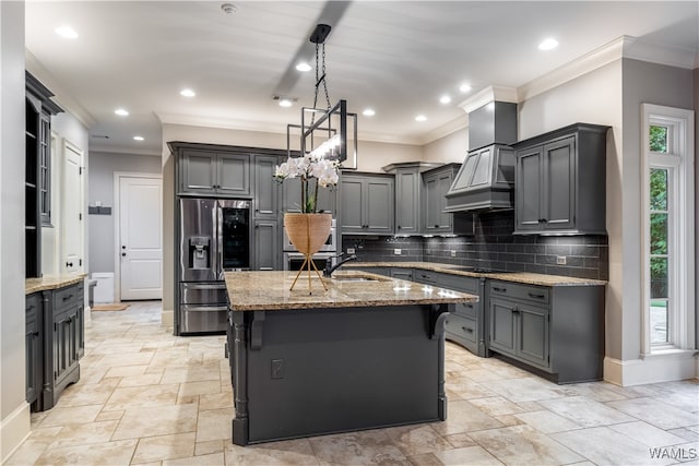 kitchen with gray cabinetry, custom exhaust hood, stainless steel refrigerator with ice dispenser, and an island with sink