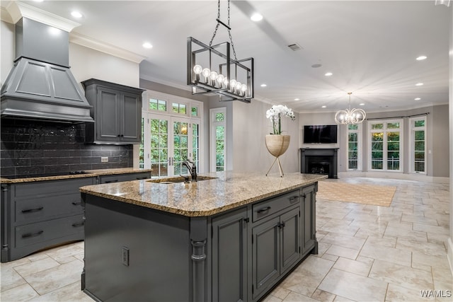 kitchen with gray cabinetry, custom exhaust hood, a kitchen island with sink, tasteful backsplash, and light stone counters