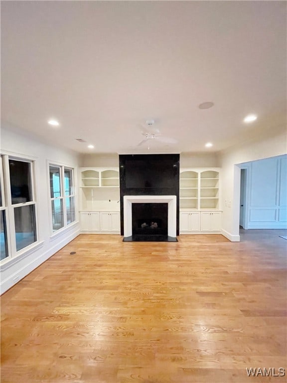 unfurnished living room with a large fireplace, built in shelves, ceiling fan, and light hardwood / wood-style flooring