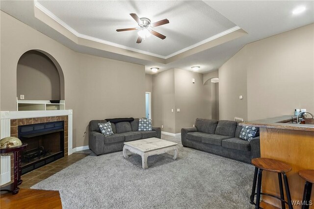 living room with a tiled fireplace, a raised ceiling, baseboards, and ornamental molding