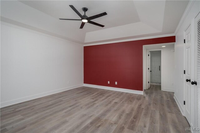 unfurnished bedroom with a tray ceiling, a ceiling fan, ornamental molding, wood finished floors, and baseboards