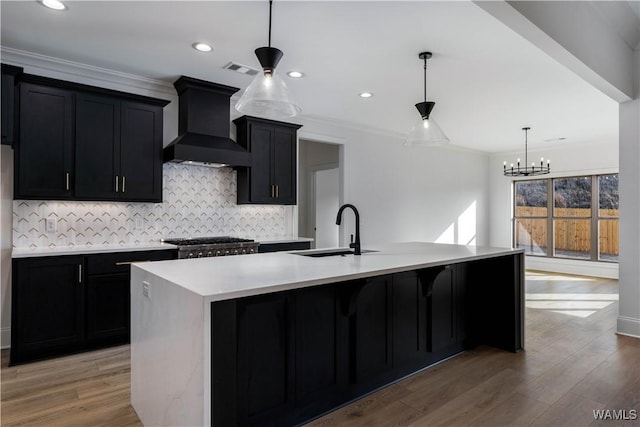 kitchen with premium range hood, wood-type flooring, sink, hanging light fixtures, and a kitchen island with sink