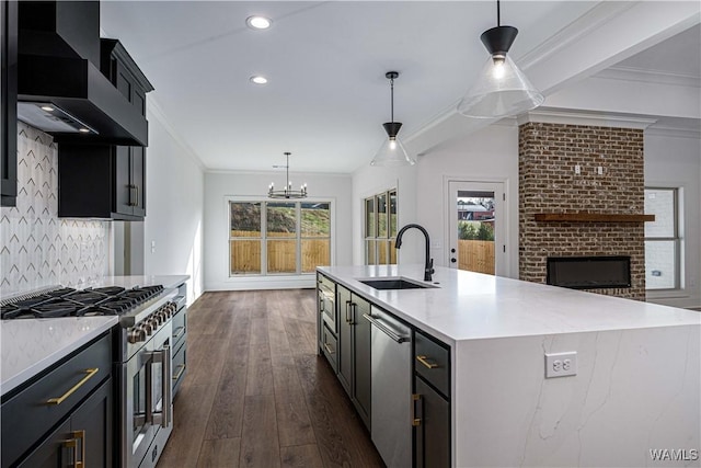 kitchen with appliances with stainless steel finishes, decorative light fixtures, sink, a kitchen island with sink, and wall chimney exhaust hood