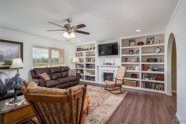 living area with arched walkways, crown molding, a premium fireplace, a ceiling fan, and wood finished floors