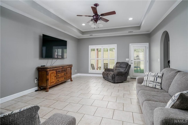 living area featuring a ceiling fan, arched walkways, a tray ceiling, baseboards, and ornamental molding