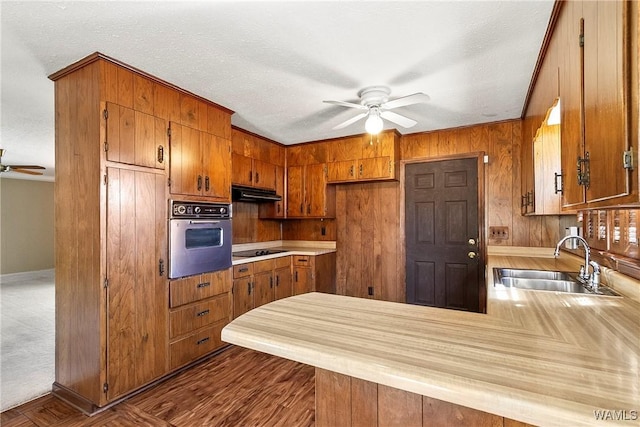 kitchen featuring a peninsula, a sink, under cabinet range hood, stainless steel oven, and brown cabinets