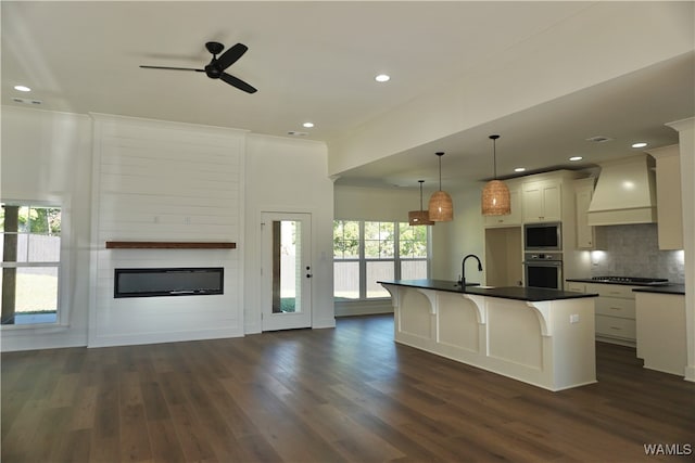 kitchen with premium range hood, stainless steel appliances, a kitchen island with sink, dark wood-type flooring, and hanging light fixtures