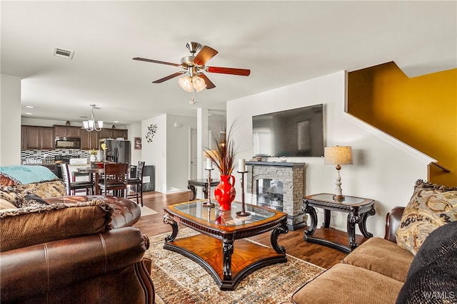 living room featuring ceiling fan with notable chandelier, a stone fireplace, and light hardwood / wood-style flooring