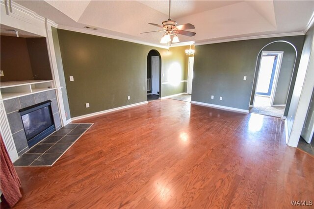 unfurnished living room featuring dark wood-type flooring, a tile fireplace, a raised ceiling, crown molding, and ceiling fan