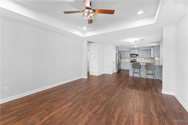 unfurnished living room featuring a tray ceiling, dark wood-style flooring, recessed lighting, ceiling fan, and baseboards