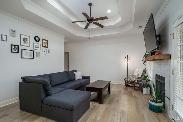 living room with crown molding, a tray ceiling, a brick fireplace, and light wood-type flooring