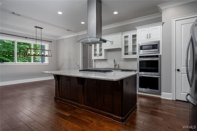 kitchen with island exhaust hood, appliances with stainless steel finishes, a center island, glass insert cabinets, and ornamental molding
