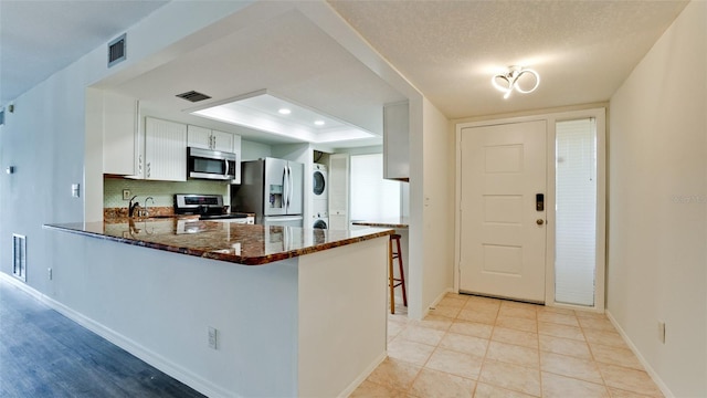 kitchen with dark stone counters, white cabinets, stainless steel appliances, kitchen peninsula, and stacked washer and dryer