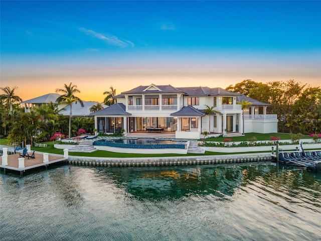 back house at dusk featuring a lawn, a swimming pool with hot tub, a balcony, and a water view