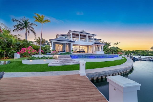 back house at dusk with a balcony, a lawn, and a water view