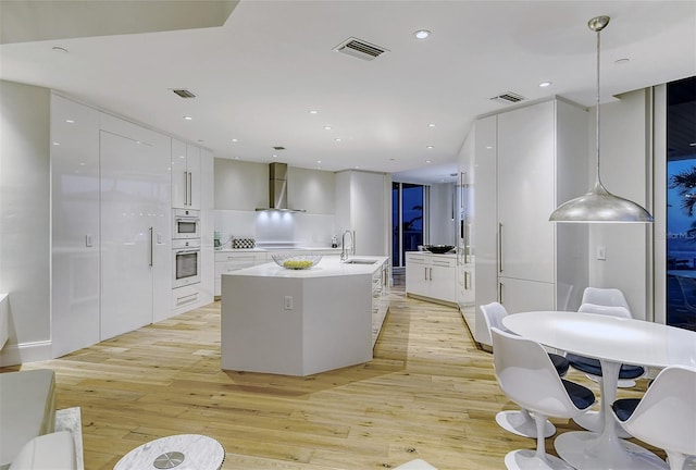 kitchen featuring white oven, a center island with sink, wall chimney exhaust hood, light wood-type flooring, and pendant lighting