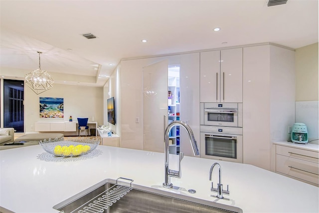 kitchen featuring double wall oven, pendant lighting, sink, an inviting chandelier, and white cabinetry