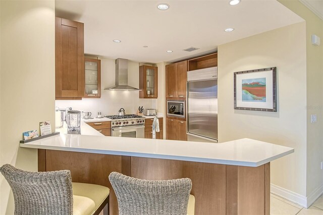 kitchen with kitchen peninsula, light tile flooring, a kitchen breakfast bar, wall chimney range hood, and built in appliances