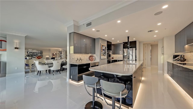 kitchen with a kitchen island with sink, gray cabinets, light tile floors, a breakfast bar, and backsplash