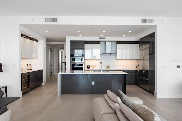 kitchen with light wood-type flooring, a kitchen island with sink, wall chimney exhaust hood, and white cabinetry