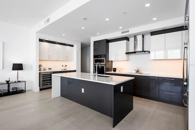 kitchen featuring wall chimney exhaust hood, an island with sink, light hardwood / wood-style floors, and white cabinetry