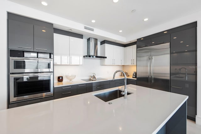 kitchen with white cabinetry, sink, an island with sink, stainless steel appliances, and wall chimney range hood