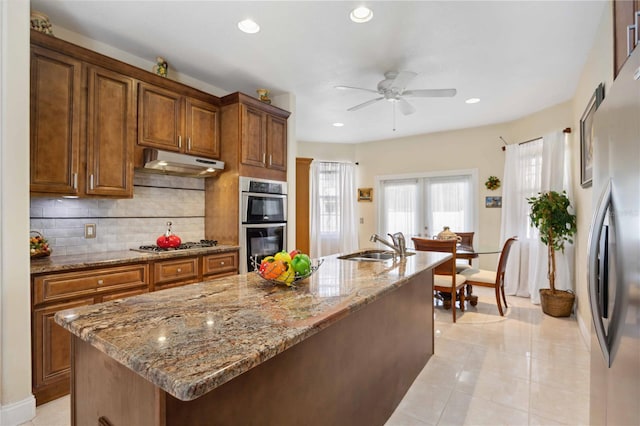 kitchen with an island with sink, ceiling fan, light stone counters, stainless steel appliances, and tasteful backsplash