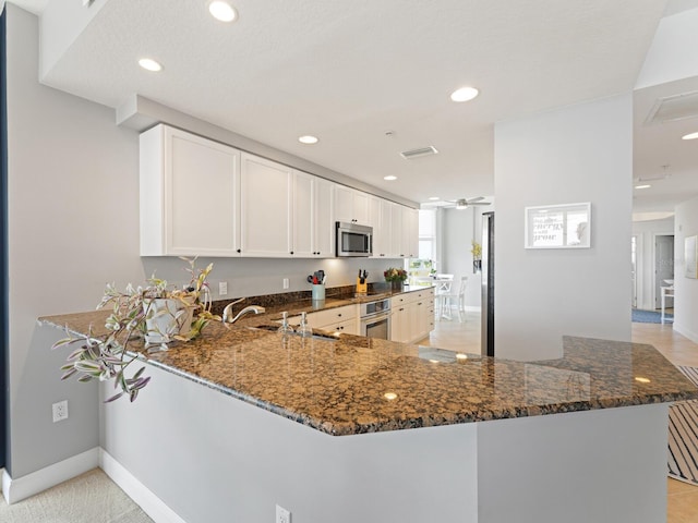 kitchen with kitchen peninsula, ceiling fan, appliances with stainless steel finishes, white cabinetry, and dark stone counters