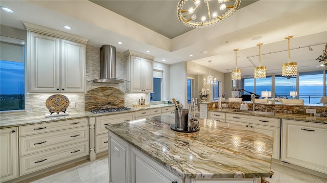 kitchen featuring wall chimney exhaust hood, a kitchen island, tasteful backsplash, an inviting chandelier, and light stone counters