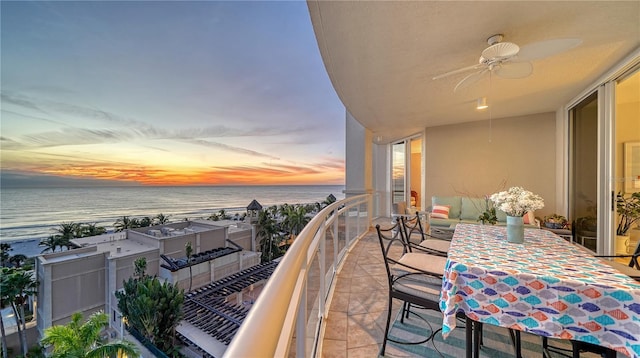 balcony at dusk with ceiling fan, a water view, and a view of the beach