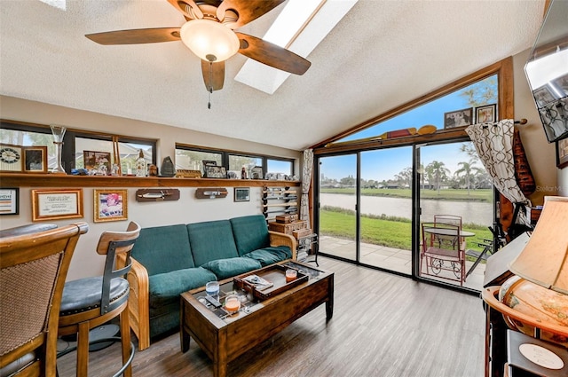 living room featuring lofted ceiling with skylight, ceiling fan, light hardwood / wood-style floors, a water view, and a textured ceiling