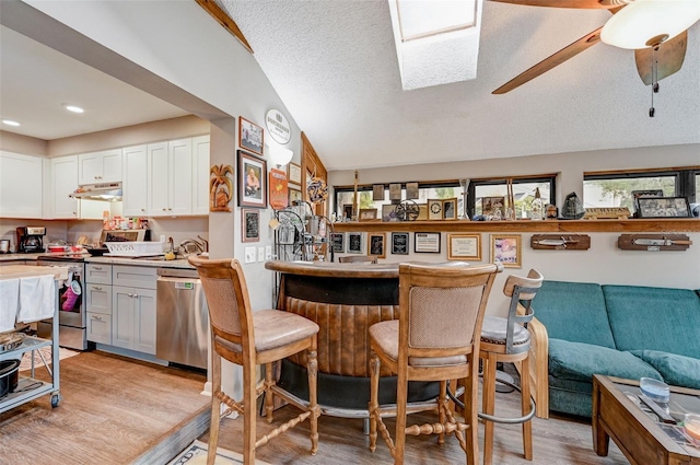 kitchen with stainless steel appliances, ceiling fan, lofted ceiling with skylight, light hardwood / wood-style flooring, and white cabinetry