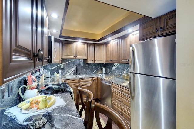 kitchen with appliances with stainless steel finishes, dark stone countertops, decorative backsplash, sink, and a tray ceiling