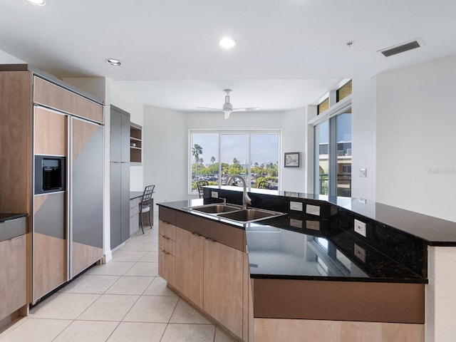 kitchen with ceiling fan, sink, a kitchen island with sink, dark stone countertops, and paneled built in fridge